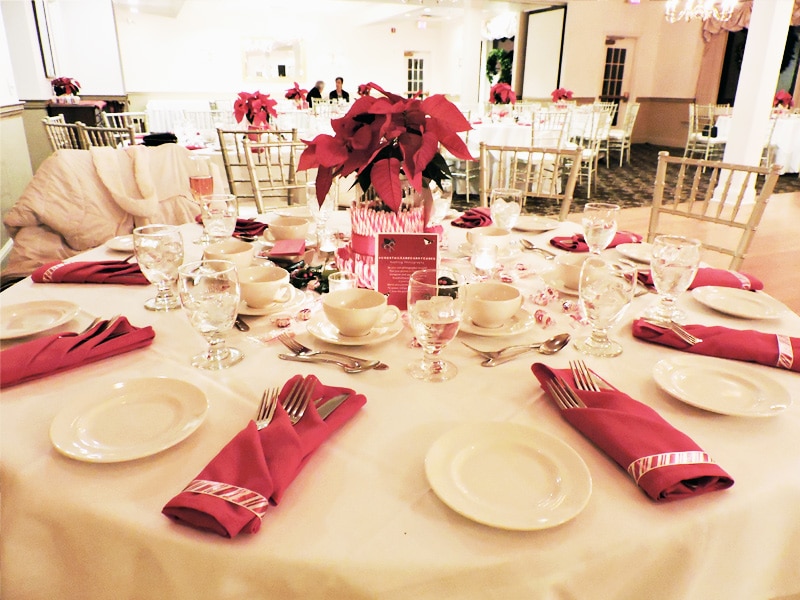 Tables in the golf club banquet hall at Copper Hill Country Club decorated with white tablecloths and red napkins and poinsettias for a holiday party.